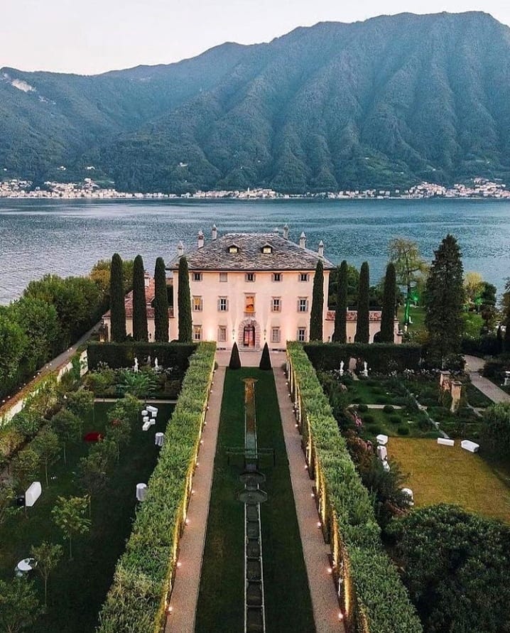 A Italian mansion with lush green gardens near a lake surrounded by mountains